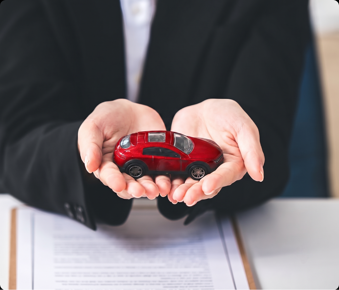 Person in a suit holding a small red model car in cupped hands above a document